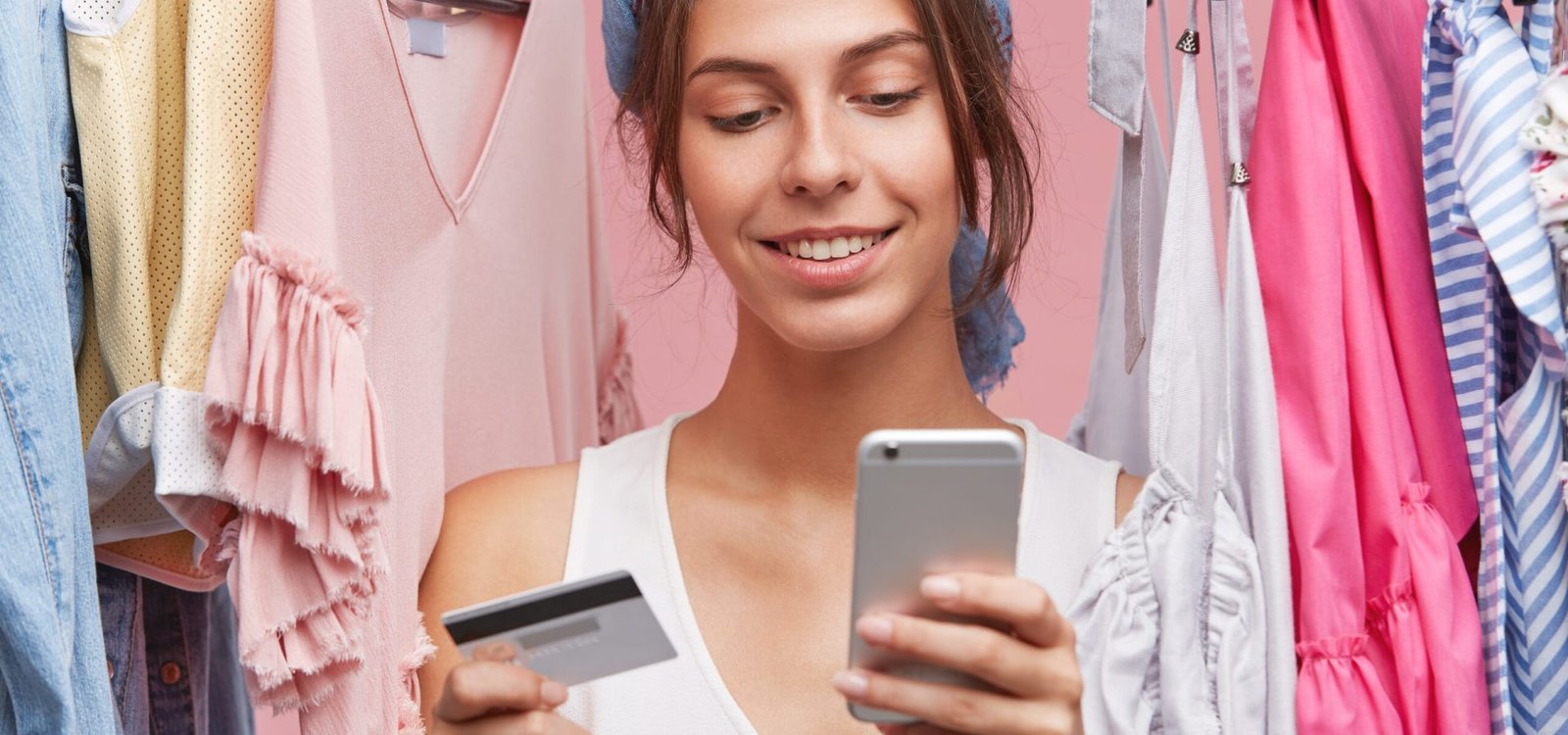 Indoor shot of beautiful woman with gentle smile, standing near variety of clothes being shopaholic, buying garment online, using smart phone and credit card. People, shopping, clothes concept Dia do Consumidor. Crédito Freepik.jpg