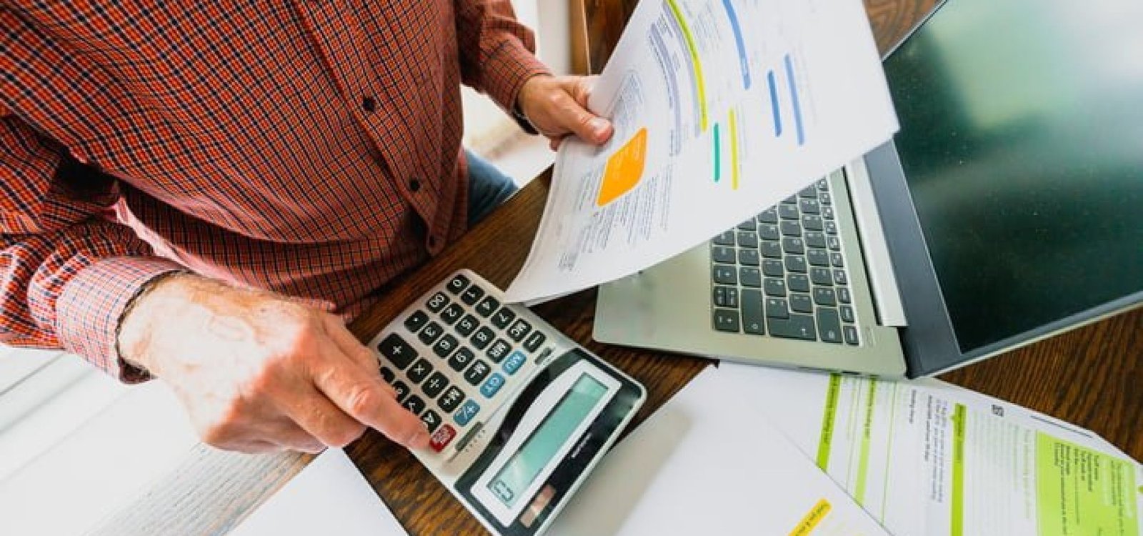 Wide angle image depicting a senior man's hand holding an energy bill while the other hand checks the numbers on a calculator. The table is strewn with documents and also a laptop. Room for copy space.