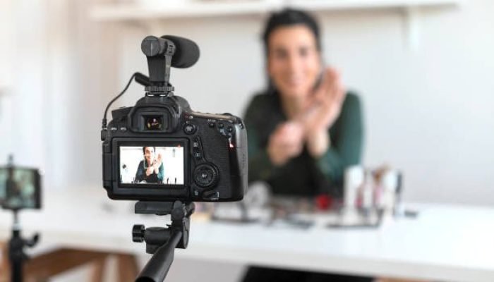 Young Woman Vlogging Herself About Beauty Products. Woman Making a Video for Her Blog on Cosmetics Using a Tripod Mounted Digital Camera. Young Female Blogger on Camera Screen Holding Cosmetics.